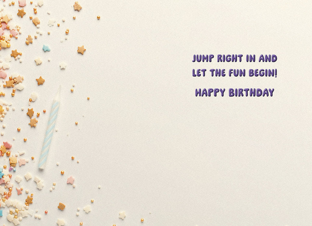 Inside of a light-colored greeting card displaying a light blue birthday candle and scattered star and pearl sprinkles, with purple text reading "JUMP RIGHT IN AND LET THE FUN BEGIN! HAPPY BIRTHDAY".