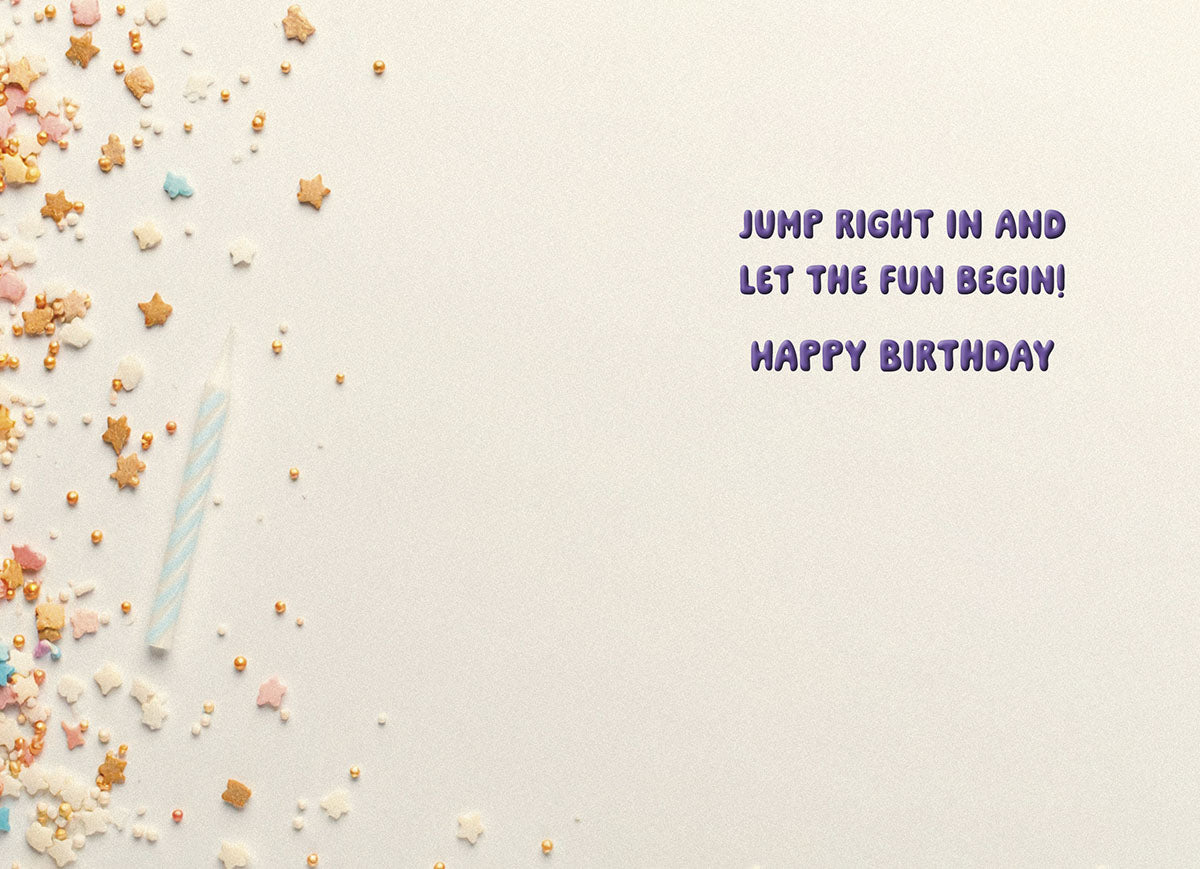 Inside of a light-colored greeting card displaying a light blue birthday candle and scattered star and pearl sprinkles, with purple text reading "JUMP RIGHT IN AND LET THE FUN BEGIN! HAPPY BIRTHDAY".