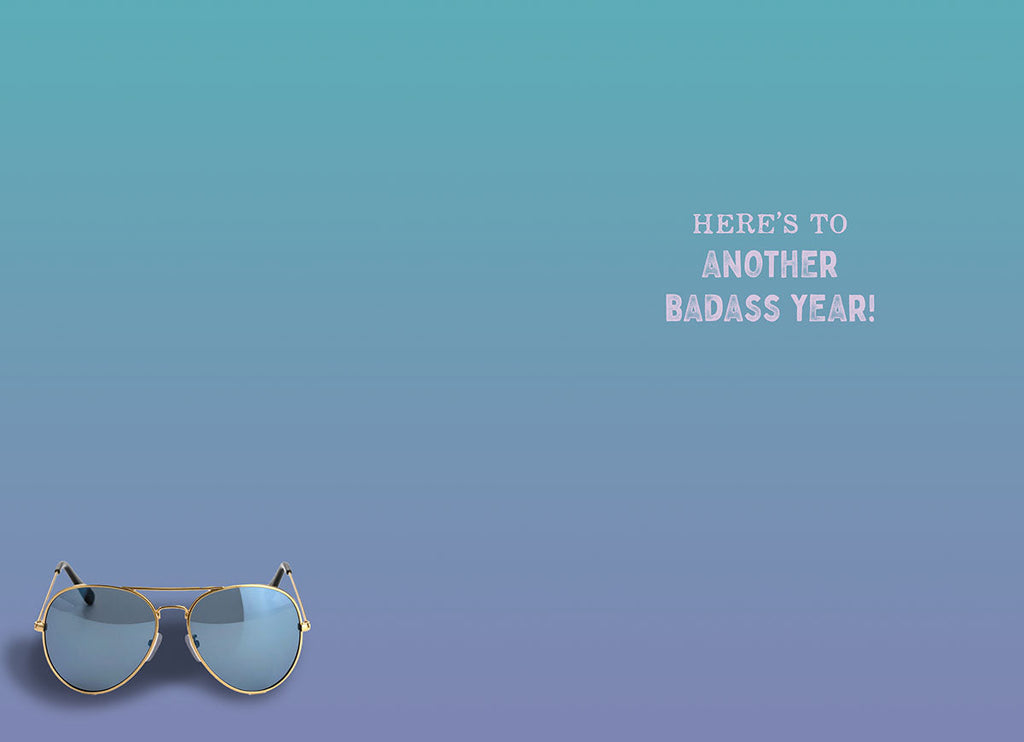 Inside view of a funny greeting card with a blue and purple gradient background, showing aviator sunglasses in the bottom left corner and the text 'HERE'S TO ANOTHER BADASS YEAR!'.