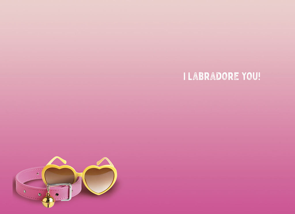 A pink dog collar with a gold bell and matching yellow heart-shaped sunglasses resting on a pink gradient background. The card's inside message "I LABRADORE YOU!" is visible.