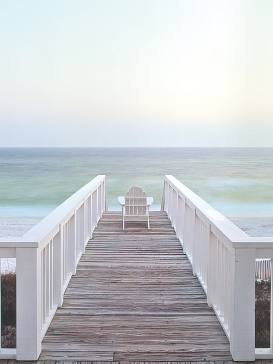 A peaceful image of a white Adirondack chair on a wooden pier extending into a calm, light blue ocean at sunset.