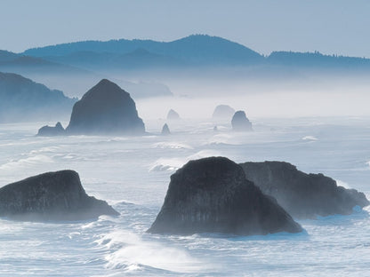 Misty coastal scene featuring prominent sea stacks rising from choppy ocean waters, with distant mountains in a blue haze.