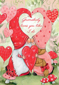 Romantic greeting card featuring two happy gnomes, one male and one female, with their pointed hats forming a heart shape, holding a large red heart. The design includes floating hearts, ladybugs, and red mushrooms against a whimsical green and pink background. Text reads: 'Gnomebody loves you like I do.'