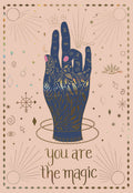 Mystical birthday greeting card featuring a dark blue hand with intricate gold foil zodiac symbols and stars on a blush pink background, with 'you are the magic' text.