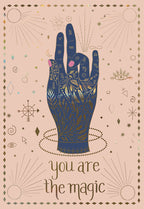 Mystical birthday greeting card featuring a dark blue hand with intricate gold foil zodiac symbols and stars on a blush pink background, with 'you are the magic' text.