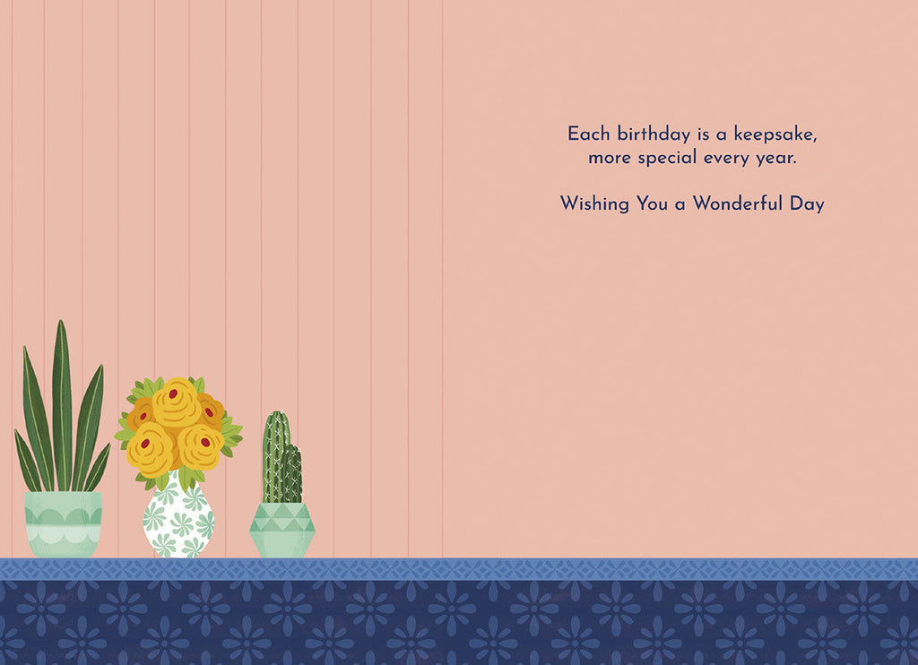 Inside of a light pink greeting card with a heartfelt message: 'Each birthday is a keepsake, more special every year. Wishing You a Wonderful Day.' Illustrated with potted plants and yellow roses along the bottom border.