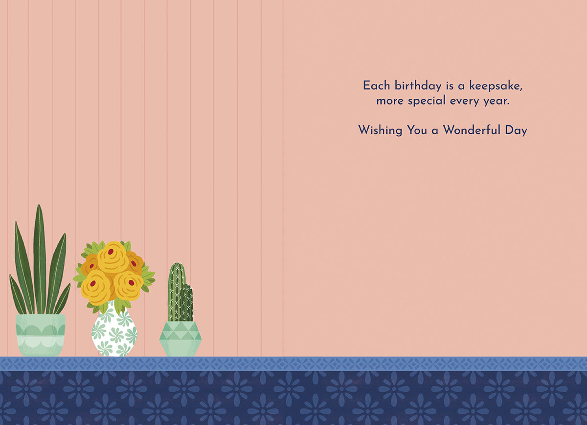 Inside of a light pink greeting card with a heartfelt message: 'Each birthday is a keepsake, more special every year. Wishing You a Wonderful Day.' Illustrated with potted plants and yellow roses along the bottom border.