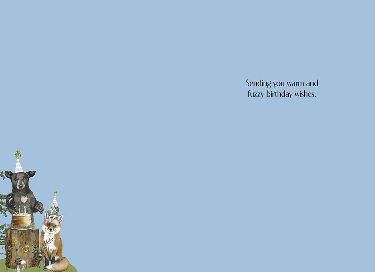 Inside of a charming greeting card featuring a small illustration of forest animals celebrating a party in the bottom left corner, with the message 'Sending you warm and fuzzy birthday wishes.' centrally placed on a light blue background.