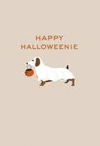 Cute dachshund dog in a white ghost costume holding an orange pumpkin candy pail, standing on a light beige background with text 'HAPPY HALLOWEENIE'. Perfect for spooky season greetings.
