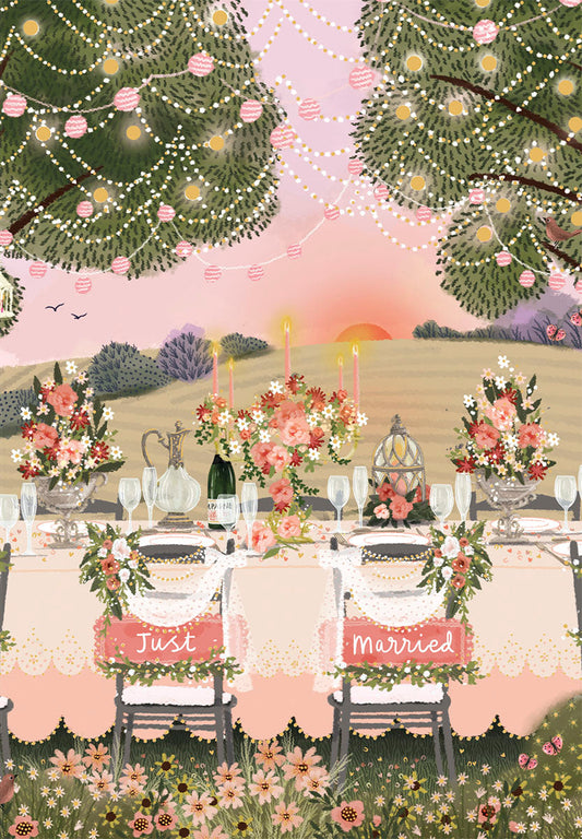 Whimsical outdoor wedding reception table at sunset with 'Just Married' chairs, floral decor, champagne, and string lights.