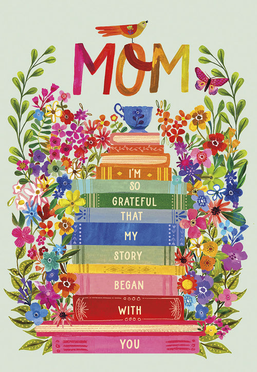 Front of a vibrant Mother's Day greeting card. It features the colorful word 'MOM' at the top, surrounded by lush green leaves and a variety of bright, whimsical flowers. A small bird perches on the 'M' of 'MOM', and a butterfly flutters nearby. Below 'MOM' is a stack of colorful books, with text across their spines reading: 'I'M SO GRATEFUL THAT MY STORY BEGAN WITH YOU'. A small blue teacup rests on the top book, adding a charming detail.