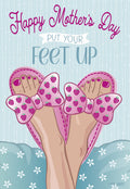 Front of a cheerful Mother's Day greeting card showing feet in pink strawberry slippers relaxing on a light blue pillow, with text 'Happy Mother's Day Put Your Feet Up' on a striped background.