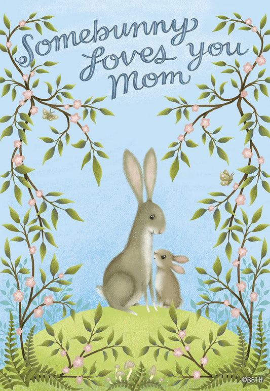 Front of a Mother's Day card with a mama bunny and baby bunny on a green hill, surrounded by flowering branches. Text reads Somebunny Loves You Mom.