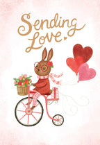 Front of a cute Valentine's Day greeting card featuring an adorable brown bunny with glasses and a scarf, riding a pink bicycle adorned with a flower basket and pulling red and pink heart balloons, with the text 'Sending Love.' against a light pink background.