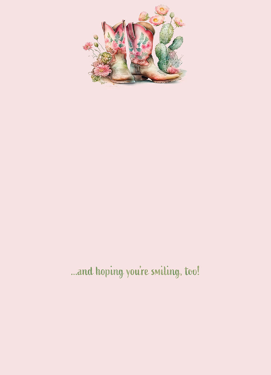 Inside of a thoughtful greeting card with a pale pink background. A small illustration of pink floral cowboy boots and desert plants is at the top, and text at the bottom reads: '...and hoping you're smiling, too!'