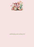 Inside of a thoughtful greeting card with a pale pink background. A small illustration of pink floral cowboy boots and desert plants is at the top, and text at the bottom reads: '...and hoping you're smiling, too!'