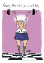 Humorous greeting card featuring an older woman with white curly hair, glasses, in a workout tank top and shorts, successfully lifting a heavy barbell above her head. The background is pink with 'Getting older makes you a hard-body.' text.