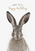 Close-up of an adorable brown hare with large ears and whiskers, looking directly ahead, against a white background with gold cursive text 'Ears to a happy birthday' above its head. Perfect for a cute greeting.