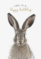 Close-up of an adorable brown hare with large ears and whiskers, looking directly ahead, against a white background with gold cursive text 'Ears to a happy birthday' above its head. Perfect for a cute greeting.