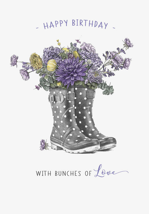 Greeting card front featuring artistic illustration of grey polka dot wellington boots overflowing with a vibrant bouquet of purple dahlias, hydrangeas, and yellow flowers, with 'Happy Birthday - with Bunches of Love' text.