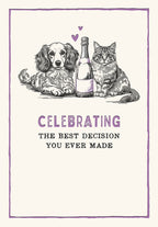 Front of a cute anniversary greeting card featuring a black and white sketch illustration of a fluffy dog and a tabby cat sitting next to a champagne bottle with two purple hearts. The text reads: 'CELEBRATING THE BEST DECISION YOU EVER MADE'.