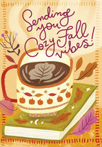 Cozy fall greeting card front featuring a warm mug of hot cocoa with leaf latte art, resting on an autumn-themed book, surrounded by fall leaves and the text 'Sending you Cozy Fall Vibes!'. Perfect for friends celebrating harvest season or leading up to a festive October.