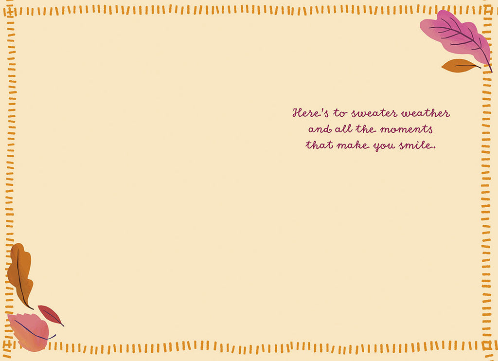 Inside of a cheerful greeting card with a light tan background and an autumn leaf border. The handwritten-style text reads 'Here's to sweater weather and all the moments that make you smile.'.