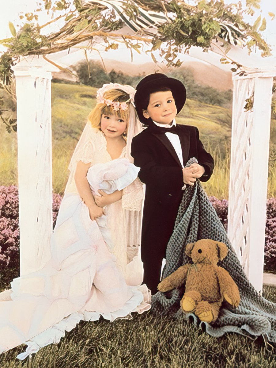 Full image of a cute young boy and girl dressed as a bride and groom, standing under a wedding arch with a teddy bear.