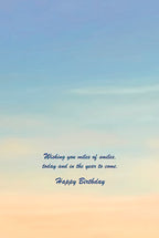 Inside of a greeting card with a serene blue and orange sky background, featuring the handwritten message 'Wishing you miles of smiles, today and in the year to come. Happy Birthday' in elegant script.