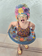 Excited young girl in a floral swim cap, green goggles, and a colorful swimsuit, holding a fun sea-creature themed pool float, perfect for a child's birthday greeting card.