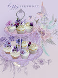 Elegant birthday greeting card featuring a two-tier stand overflowing with white-frosted cupcakes, adorned with delicate purple and pink floral accents, on a lavender background with intricate botanical illustrations.