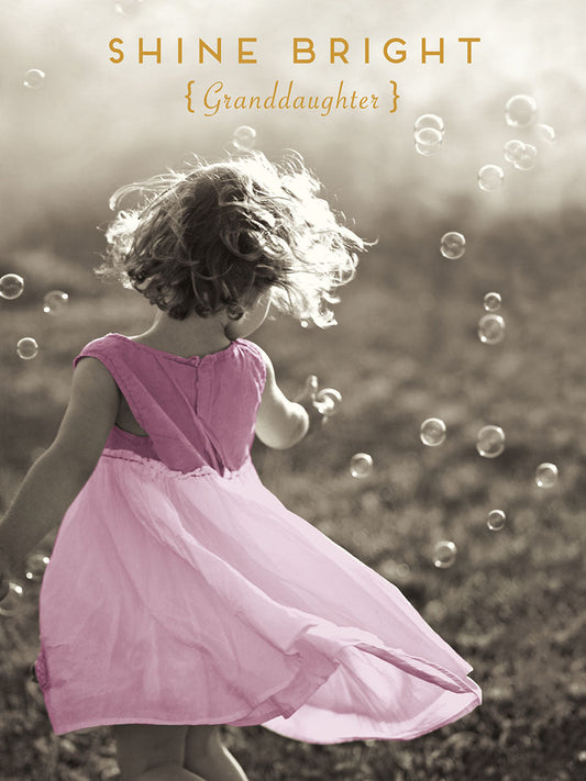 Rear view of a happy young girl in a flowing pink dress, playing with bubbles in a sunlit field. Text: "SHINE BRIGHT {Granddaughter}".