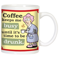 Front view of a white ceramic coffee mug featuring a funny cartoon of an older woman in a bathrobe with pink hair, holding a steaming coffee cup. Text reads: 'Coffee keeps me busy until it's time to be drunk.' on a yellow and green background.