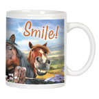 Close-up of a white ceramic mug featuring a smiling brown horse and a dark horse peeking from behind a wooden fence, with "Smile!" text and a sunny landscape background.