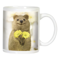 White ceramic coffee mug featuring a smiling quokka holding two cheerful yellow daisy-like flowers on a bright, blurry background.