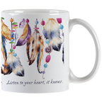 Close-up of a white ceramic mug, showing the right side with a handle. The mug features a vibrant design of hanging feathers in various earthy and pastel colors, interspersed with colorful beads. Part of the quote 'Listen to your heart, it knows.' is visible in elegant script below the design.
