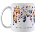 White ceramic coffee mug viewed from the left side with its handle. The mug is adorned with an intricate pattern of watercolor feathers and bright beads, suggesting a spiritual or bohemian theme. The inspirational message 'Listen to the wind, it talks.' is elegantly scripted on the mug's lower portion.
