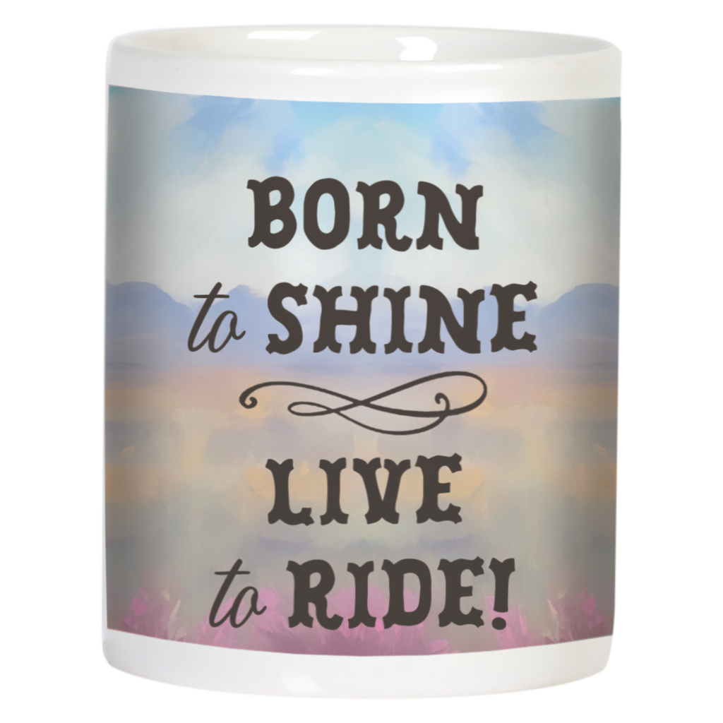 Back view of a white ceramic mug displaying the inspirational text "BORN to SHINE LIVE to RIDE!" in dark brown script over a soft watercolor background with distant mountains and a serene sunset sky.