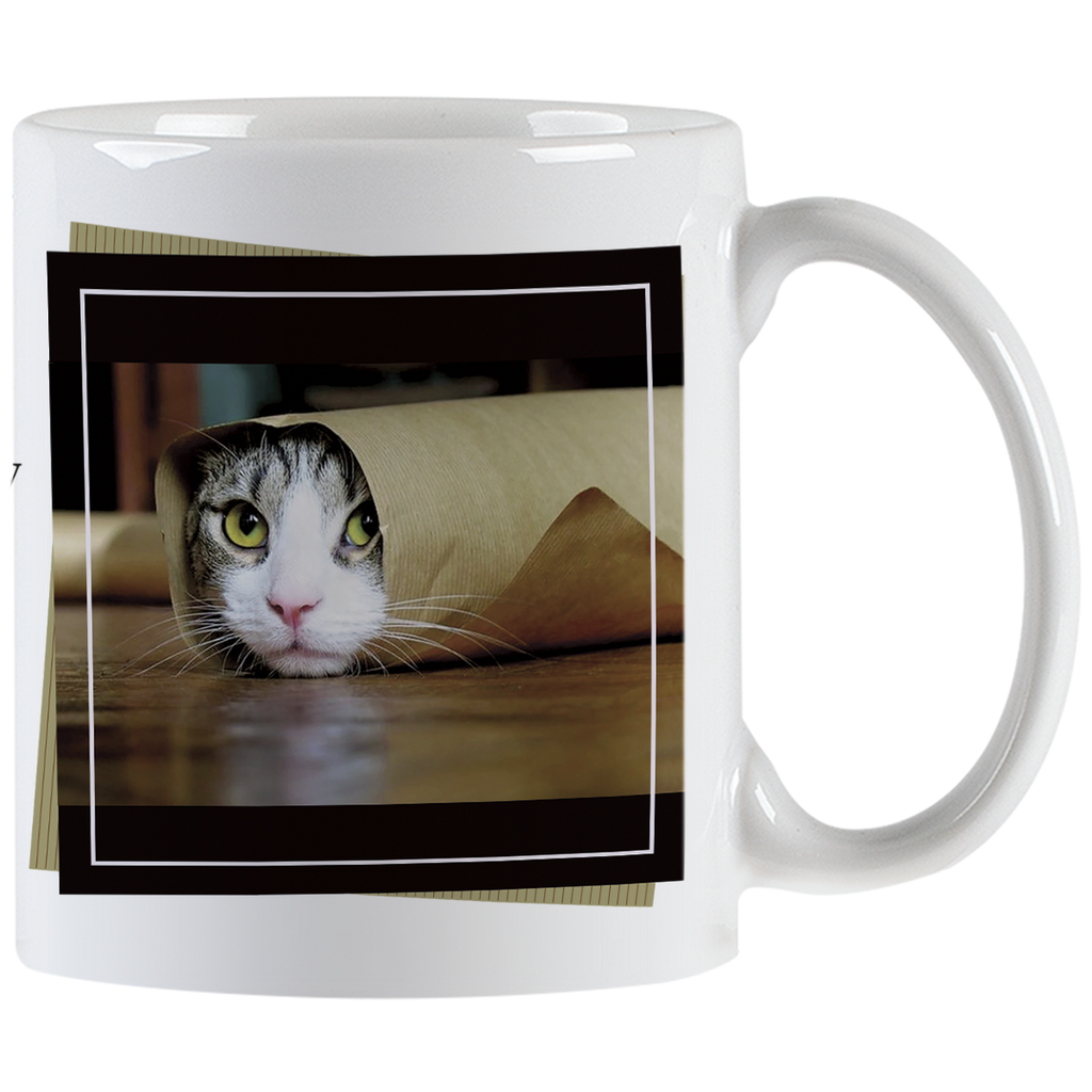 Close-up of a white ceramic mug featuring a tabby cat peeking out from a rolled-up brown paper bag with bright green eyes, framed by a black border.