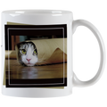 Close-up of a white ceramic mug featuring a tabby cat peeking out from a rolled-up brown paper bag with bright green eyes, framed by a black border.