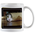 Close-up of a white ceramic mug featuring a tabby cat peeking out from a rolled-up brown paper bag with bright green eyes, framed by a black border.