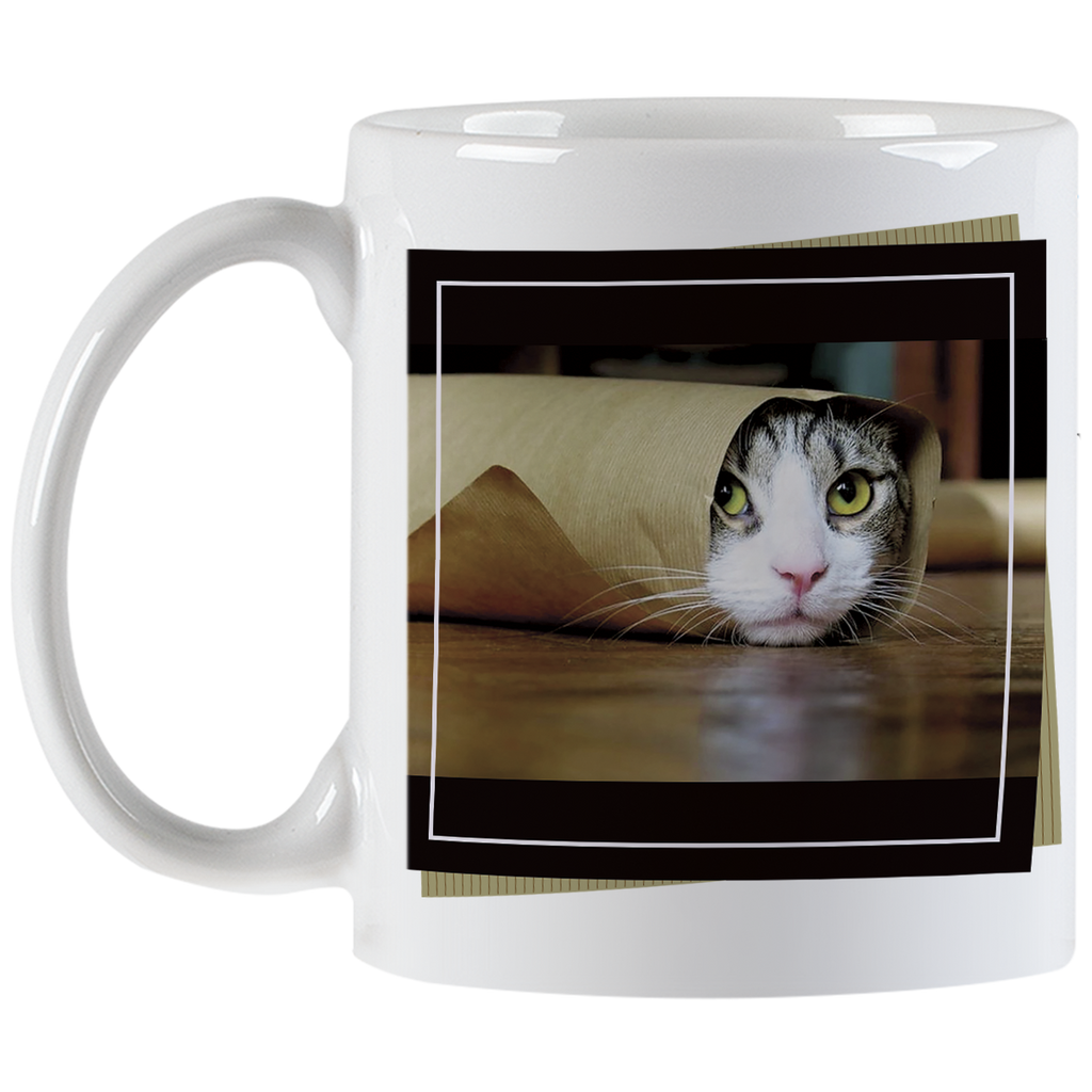Angled view of a white ceramic mug showing a tabby cat with striking green eyes peeking from a brown paper bag, against a blurred dark background, framed by a black border.