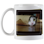 Angled view of a white ceramic mug showing a tabby cat with striking green eyes peeking from a brown paper bag, against a blurred dark background, framed by a black border.
