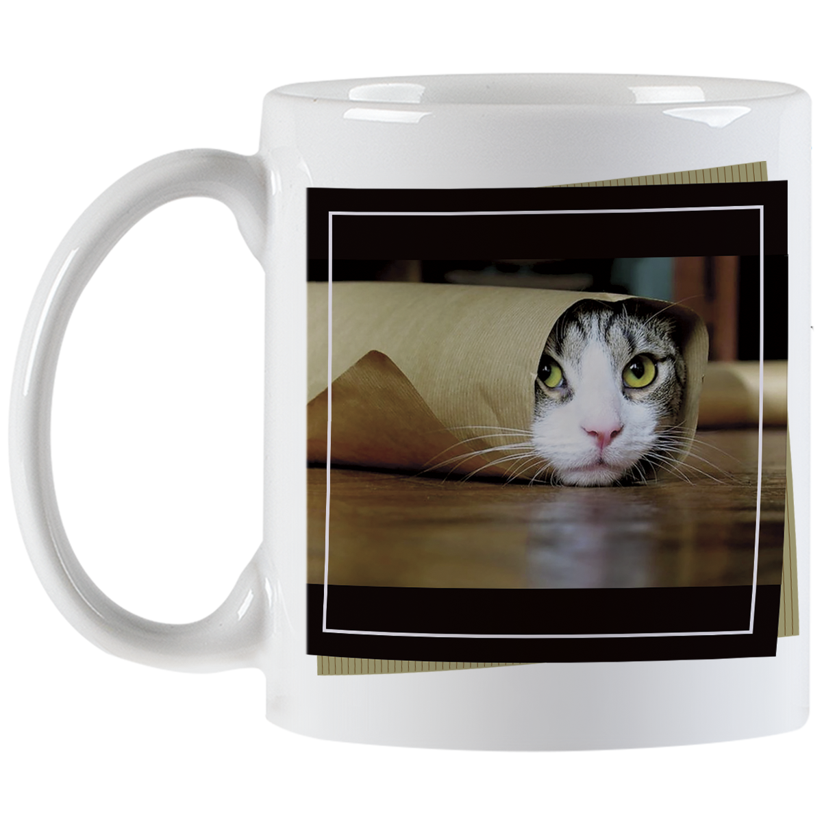 Angled view of a white ceramic mug showing a tabby cat with striking green eyes peeking from a brown paper bag, against a blurred dark background, framed by a black border.