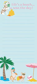 Magnetic list pad with a beach theme. Top: A Dalmatian dog wearing a straw hat and floral lei, holding a green bucket, stands beside a sandcastle and a red crab. The phrase 'life's a beach... seas the day!' is in pink text. Bottom: A palm tree, a Shiba Inu dog in a blue cap holding an ice cream, and a Corgi dog in sunglasses and an orange life vest. A pink and white striped beach umbrella and a beach ball are also visible. The background is light blue with ruled lines for writing.