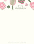 An SEO-optimized alt description for a light beige notepad. The top border features pink and green pickleball paddles, some with floral daisy patterns, others with strawberry designs. Dotted pickleballs are interspersed among the paddles. The central text reads 'In my Pickleball Era' in a decorative script. This design is perfect for pickleball players, fans, and anyone looking for fun, themed stationery.