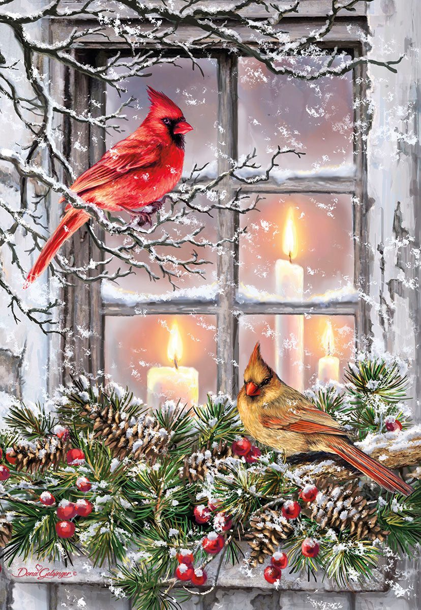 Front of a festive holiday greeting card featuring a vibrant red male cardinal and a brown female cardinal perched on snow-covered branches outside a window. Warm candlelight glows from inside, surrounded by pinecones, evergreen boughs, and red berries, evoking a cozy winter scene.