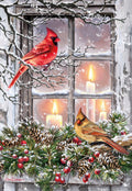 Front of a festive holiday greeting card featuring a vibrant red male cardinal and a brown female cardinal perched on snow-covered branches outside a window. Warm candlelight glows from inside, surrounded by pinecones, evergreen boughs, and red berries, evoking a cozy winter scene.