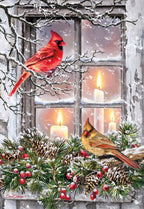 Front of a festive holiday greeting card featuring a vibrant red male cardinal and a brown female cardinal perched on snow-covered branches outside a window. Warm candlelight glows from inside, surrounded by pinecones, evergreen boughs, and red berries, evoking a cozy winter scene.