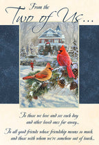 Front of a winter greeting card featuring two vibrant red cardinals perched on a snow-dusted pine branch in front of a festive, elegant Victorian house, with 'From the Two of Us...' script.